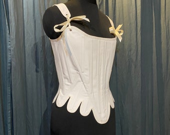 May include: A white cotton corset with a scalloped bottom and ribbon ties. The corset is on a black dress form.