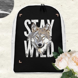 Könnte beinhalten: Schwarzer Rucksack mit einer weißen Wolfsillustration und dem Text "STAY WILD" in weißen Buchstaben. Der Rucksack hat einen weißen Rand um die Kanten.