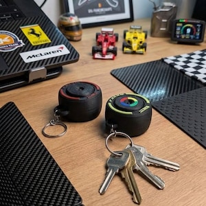 Può includere: Due portachiavi neri circolari a forma di pneumatici da corsa. Un portachiavi presenta un anello rosso sulla superficie superiore con la parola Ferrari, mentre l'altro ha un cerchio centrale colorato e un bordo verde chiaro. Entrambi includono anelli.