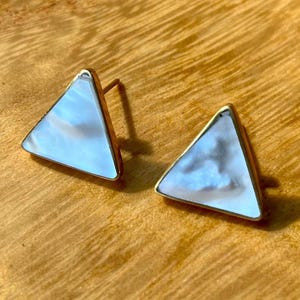 Op de afbeelding: Een paar goudkleurige driehoekige oorstekers met een parelmoer, iriserende witte inleg. De oorbellen zijn geplaatst op een warm houten oppervlak, dat hun geometrische ontwerp en subtiele kleurvariaties benadrukt.