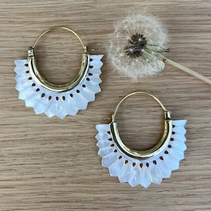 Op de afbeelding: Een paar goudkleurige oorringen met een wit, geschulpte, waaiervormig ontwerp. De oorbellen hebben een gepolijste afwerking en worden op een houten oppervlak gepresenteerd. Een modeaccessoire.