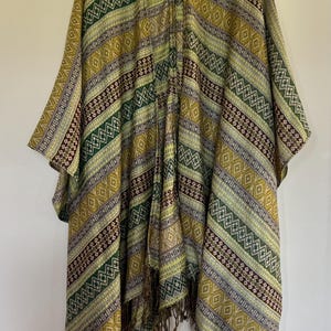 May include: A patterned poncho in shades of green, gold, and brown hangs on a wooden hanger. The poncho features a geometric design with stripes and diamond shapes. The bottom edge has a fringe detail. The garment is made of a soft, woven fabric.
