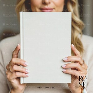 A lifestyle photograph of a woman softly smiling while holding a blank, cream-colored A5 hardcover book directly in front of her. She is holding the book with both hands, showcasing a delicate rose gold watch and rings. The lighting is soft and natural, creating a feminine, high-end aesthetic.