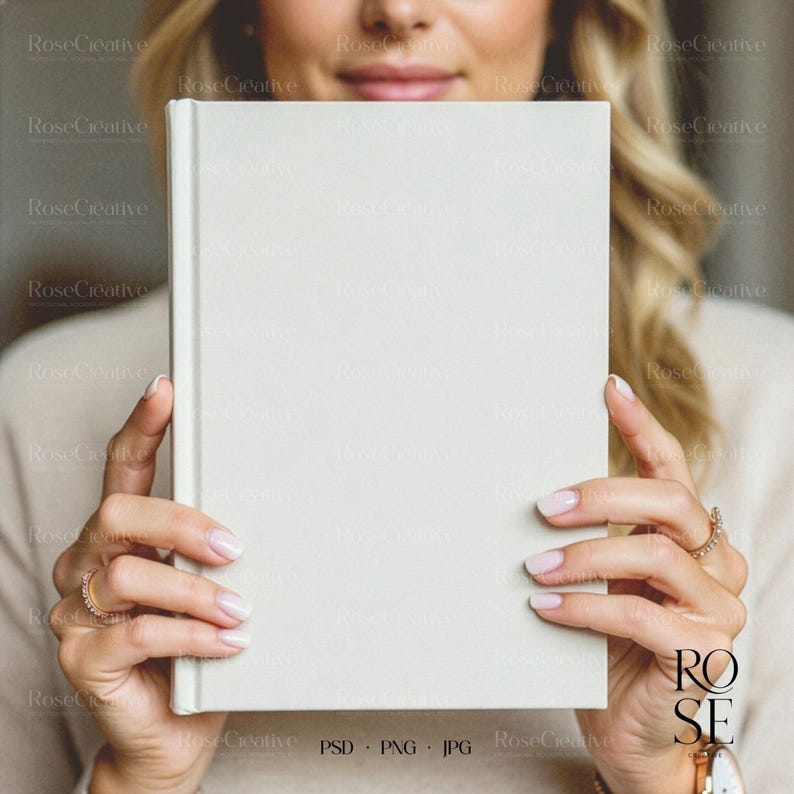 A lifestyle photograph of a woman softly smiling while holding a blank, cream-colored A5 hardcover book directly in front of her. She is holding the book with both hands, showcasing a delicate rose gold watch and rings. The lighting is soft and natural, creating a feminine, high-end aesthetic.