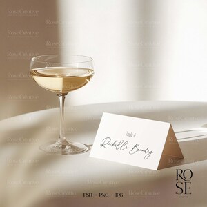 May include: A champagne coupe glass filled with a golden liquid, next to a folded white place card with the name "Rochelle Beandry" and "Table 4" written in elegant script. The image has a soft, neutral background.