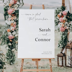 May include: A white wedding welcome sign with black calligraphy text that reads "We're so glad that you're here" and "Sarah and Connor" with the date "June 16, 2023" and location "Bella Beach, CA". The sign is on an easel in front of a floral archway.