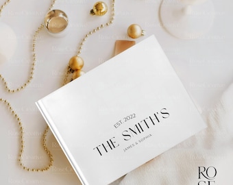 May include: A white book with the text "EST. 2022 THE SMITH'S JAMES & SOPHIA" in black. The book is surrounded by gold ornaments and a gold beaded garland. The book is on a white surface.