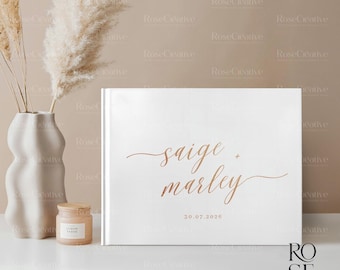 May include: A white wedding guest book with the names "saige + marley" in a copper script font and the date "30.07.2026". The book is on a white surface, next to a vase with dried flowers and a small candle.