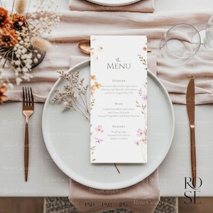 May include: A wedding menu card with floral accents, featuring the text "the MENU". The card is placed on a white plate, with a gold fork and knife. The table setting includes a floral arrangement and glassware.