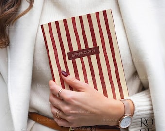 May include: A notebook with a cream and red striped cover is held by a person. The word "SERENDIPITY" is printed on a dark red rectangular label. The person is wearing a white sweater, coat, and a watch with a rose gold band.