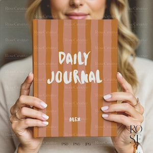 A lifestyle photograph of a woman softly smiling while holding a blank, cream-colored A5 hardcover book directly in front of her. She is holding the book with both hands, showcasing a delicate rose gold watch and rings. The lighting is soft and natural, creating a feminine, high-end aesthetic.