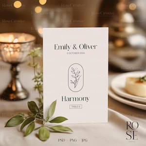 May include: A white table number card with black text. The card reads "Emily & Oliver" and "5 October 2024". Below the text is a simple line drawing of a flower in a circle. The text "Harmony" and "Table 6" are below the flower.