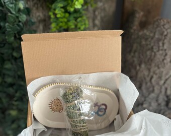 May include: A beige cardboard box contains a white oval dish with gold trim and a bundle of dried herbs wrapped in twine. The dish has colorful sun and abstract designs. The box is lined with white tissue paper.