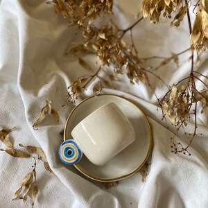 May include: A white ceramic coffee cup with a gold rim and a blue and white evil eye charm sits on a white saucer. The cup and saucer are on a white fabric background.
