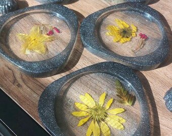 Op de afbeelding: Set van vijf grijze harsonderzetters met een transparante epoxytop. Elke onderzetter heeft een ander geperst bloemenmotief met gele bloemen en rode accenten.