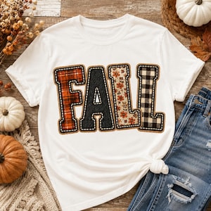 May include: A white short-sleeve t-shirt featuring the word FALL in large block letters. Each letter has a distinct pattern: the F is red plaid, the A is black herringbone, the L is a floral print with orange flowers, and the L is black and white buffalo check.