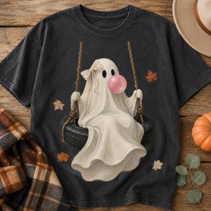 May include: A charcoal gray t-shirt featuring a graphic of a white ghost with a beige bow on its head, blowing a pink bubble gum bubble while sitting on a black tire swing suspended by ropes, surrounded by scattered orange and brown autumn leaves.