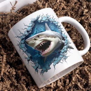 May include: White ceramic mug with a 3D image of a great white shark breaking through a wall. The shark has a blue background and is shown with its mouth open.