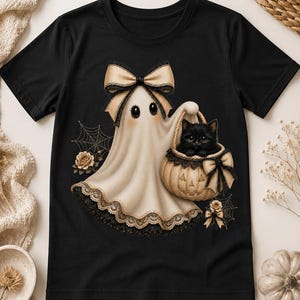 May include: A black t-shirt featuring a graphic of a cream-colored ghost wearing a large bow, holding a pumpkin-shaped basket with a black kitten inside. The ghost has lace trim at the bottom, and the design includes spiderwebs and roses.