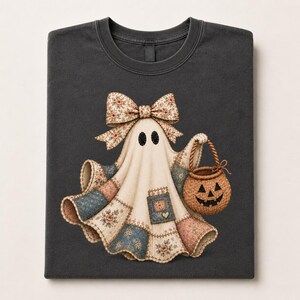 Op de afbeelding: Een antracietgrijs t-shirt met een afbeelding van een geest gemaakt van patchworkstof. De geest heeft een grote bloemrijke strik op het hoofd en draagt een gevlochten bruine pompoenmand. Het patchworkontwerp bevat blauwe, roze en crèmekleurige bloemenpatronen.