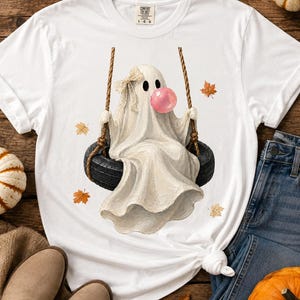 May include: A white t-shirt featuring a graphic of a ghost sitting on a black tyre swing. The ghost wears a small beige bow on its head and is blowing a large pink bubble. Several orange and yellow autumn leaves are scattered around the ghost.