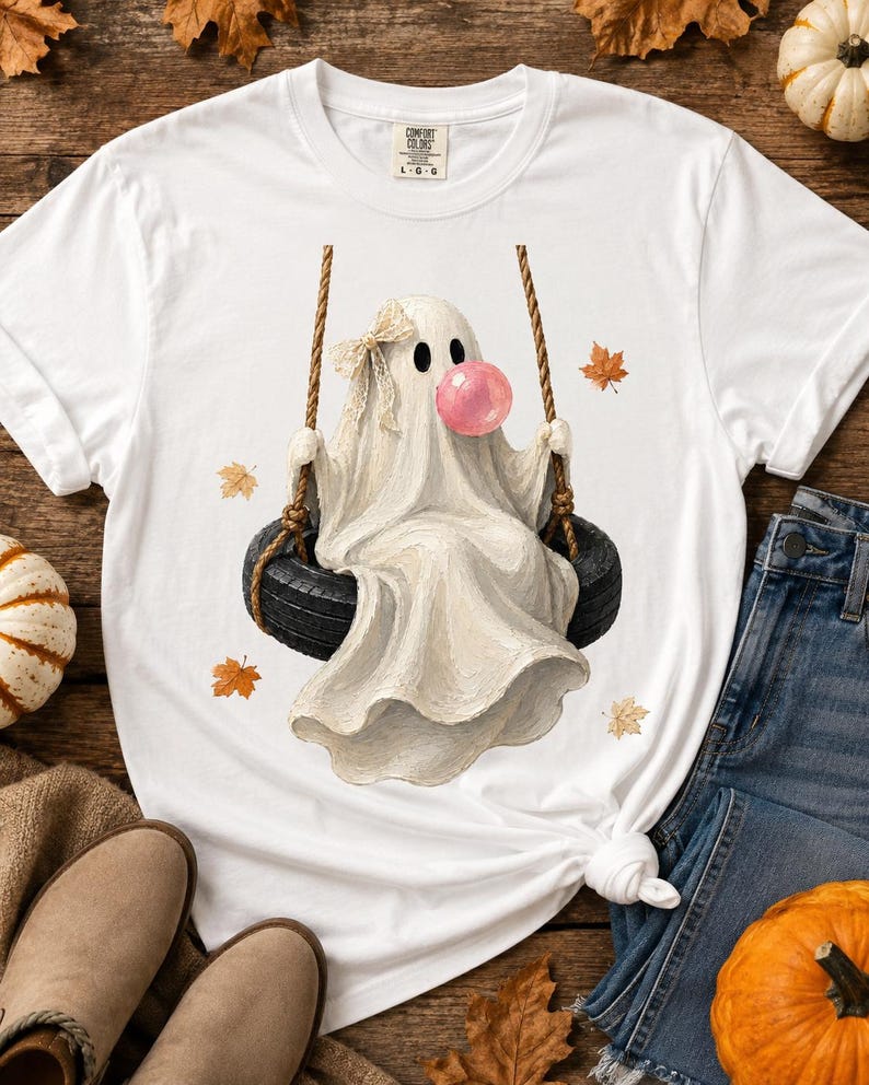 May include: A white t-shirt featuring a graphic of a ghost sitting on a black tyre swing. The ghost wears a small beige bow on its head and is blowing a large pink bubble. Several orange and yellow autumn leaves are scattered around the ghost.