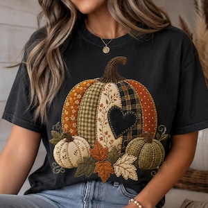 May include: A charcoal gray short-sleeve t-shirt featuring a large central graphic of a patchwork-style pumpkin. The pumpkin design incorporates various patterns including plaid, floral, and polka dots in orange, green, and cream, with a black heart.