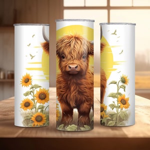 Puede incluir: Tres vasos blancos de acero inoxidable. Cada vaso presenta una ilustraci&oacute;n de una vaca de las Tierras Altas contra un sol amarillo, con girasoles y hierba. Los vasos est&aacute;n dise&ntilde;ados para bebidas fr&iacute;as.