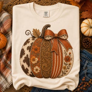 May include: A cream-colored crewneck sweatshirt featuring a large graphic of a pumpkin. The pumpkin is divided into vertical sections with cow print, orange floral patterns, brown scrollwork, and orange stripes, topped with a large orange bow.