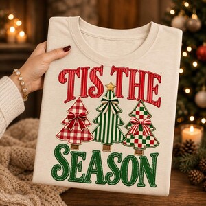 May include: A beige t-shirt featuring the red text 'TIS THE' and green text 'SEASON'. Between the text are three stylized Christmas trees: one with a red and white gingham pattern, one with green and white stripes, and one with a red and green checkerboard pattern, each adorned with a small bow.