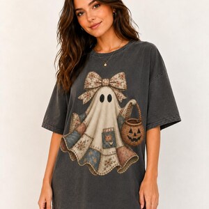 Puede incluir: Una camiseta de manga corta gris carbón con un gráfico de un fantasma que lleva un gran lazo con estampado floral en la cabeza. El fantasma está cubierto con una tela estilo patchwork con varios patrones de flores y estrellas y lleva un cubo de calabaza.