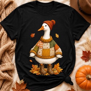 Puede incluir: Una camiseta negra de manga corta con el gráfico de un ganso blanco que lleva un gorro de punto naranja, un suéter de retazos multicolor y botas de goma marrones, de pie entre hojas de otoño naranjas y amarillas esparcidas.