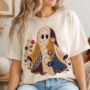 May include: A cream-colored short-sleeve t-shirt featuring a central graphic of a ghost composed of various fabric patterns including floral, plaid, and quilted textures in shades of mustard yellow, navy blue, purple, and white with small flowers.
