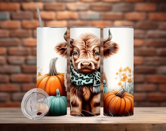 May include: A white tumbler with a brown and white highland cow wearing a blue and brown scarf. The tumbler is surrounded by pumpkins and fall foliage.