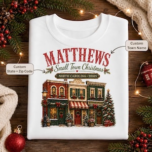 May include: A white sweatshirt featuring a graphic of a small town street with three buildings decorated for Christmas. The text reads MATTHEWS Small Town Christmas and NORTH CAROLINA 28105. The design includes a street lamp and a Christmas tree.