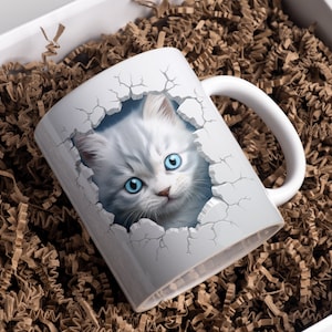 Op de afbeelding: Witte keramische mok met een 3D-afbeelding van een wit kitten met blauwe ogen dat uit een gebarsten muur kijkt.