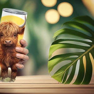 Puede incluir: Un vaso de acero inoxidable con una ilustraci&oacute;n de una vaca de las Tierras Altas. El dise&ntilde;o incluye un fondo amarillo con un girasol y hierba. La vaca tiene pelaje marr&oacute;n y cuernos blancos. El vaso es sostenido por una mano.