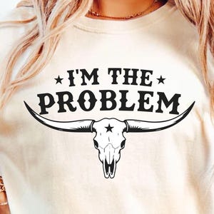 May include: Cream-colored t-shirt with the text "I'M THE PROBLEM" above a black bull skull graphic. The bull skull has long horns and a star on the forehead. The shirt is a casual, short-sleeved style.