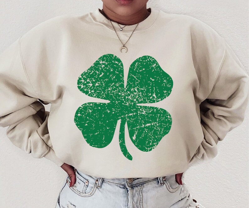 Peut inclure: Un sweat-shirt beige clair avec un graphique de tr&egrave;fle &agrave; quatre feuilles vert. Le tr&egrave;fle a un aspect vieilli et d&eacute;lav&eacute;.