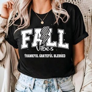 May include: Black t-shirt with white block letters spelling "FALL" and "Vibes". A checkered lightning bolt graphic is in the center. The words "THANKFUL GRATEFUL BLESSED" are below the word "Vibes".