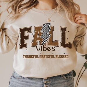 May include: A cream-coloured sweatshirt featuring the text "FALL Vibes" in brown block letters and a checkered lightning bolt graphic. The words "THANKFUL GRATEFUL BLESSED" are below. The sweatshirt is worn with blue jeans.