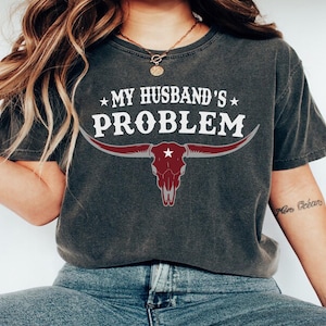 Può includere: T-shirt grigio scuro con la scritta bianca "MY HUSBAND'S PROBLEM" sopra una grafica di teschio di toro bordeaux. La maglietta ha maniche corte e girocollo. La maglietta è indossata da una persona.