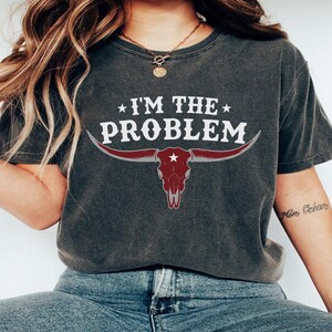May include: Dark gray t-shirt with the text "I'M THE PROBLEM" in white, above a maroon bull skull graphic. The bull skull has long horns and a star in the center. The shirt is a casual style.