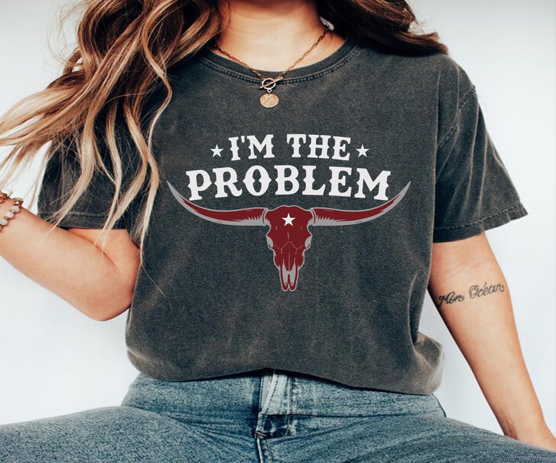 May include: Dark gray t-shirt with the text "I'M THE PROBLEM" in white, above a maroon bull skull graphic. The bull skull has long horns and a star in the center. The shirt is a casual style.