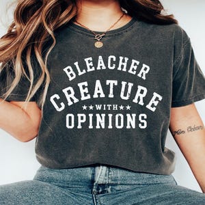 May include: Dark gray t-shirt with the text "BLEACHER CREATURE WITH OPINIONS" in white block letters. The shirt has a crew neck and short sleeves. The shirt is worn with blue jeans.