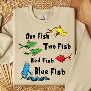 May include: A beige sweatshirt with a graphic of four fish and the text "One Fish Two Fish Red Fish Blue Fish".