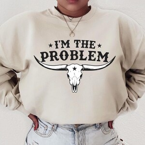 May include: Cream-colored sweatshirt with the text "I'M THE PROBLEM" above a black and white longhorn skull graphic. The sweatshirt has a crew neck and long sleeves, and is made of a soft material.
