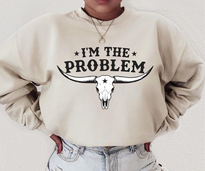 May include: Cream-colored sweatshirt with the text "I'M THE PROBLEM" above a black and white longhorn skull graphic. The sweatshirt has a crew neck and long sleeves, and is made of a soft material.