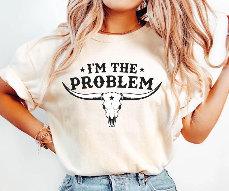 May include: Cream-colored t-shirt with the text "I'M THE PROBLEM" above a black bull skull graphic. The bull skull has long horns and a star on the forehead. The shirt is a casual, short-sleeved style.