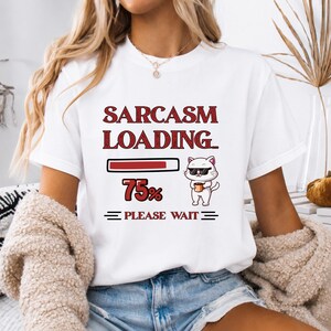 May include: A white short-sleeve t-shirt featuring a red graphic that reads SARCASM LOADING... with a progress bar at 75 percent. Next to the text is a cartoon white cat wearing black sunglasses and holding a small brown mug. Below, it says PLEASE WAIT.
